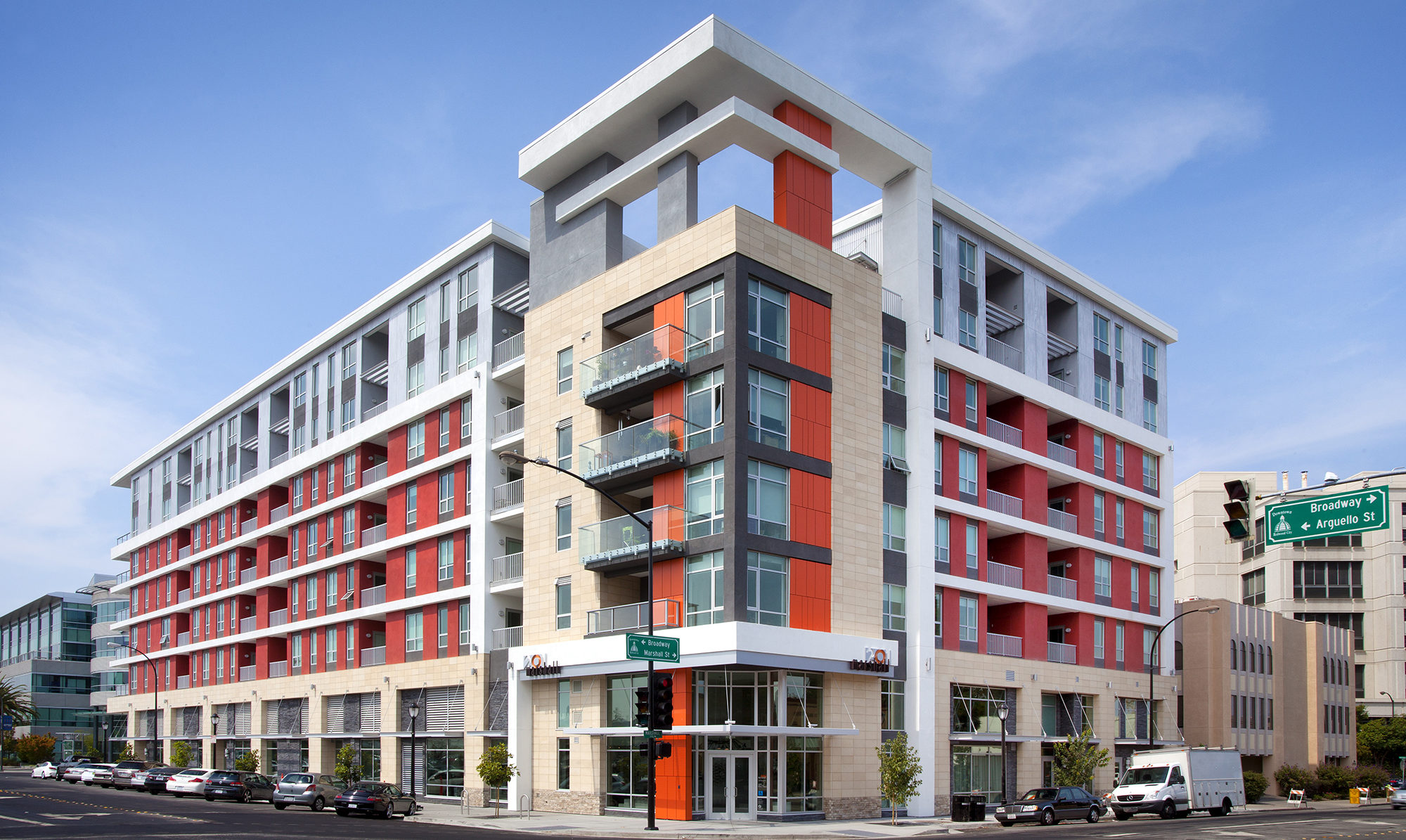 Front of podium building from street view with a lot of windows and balconies, along with red accent panels.