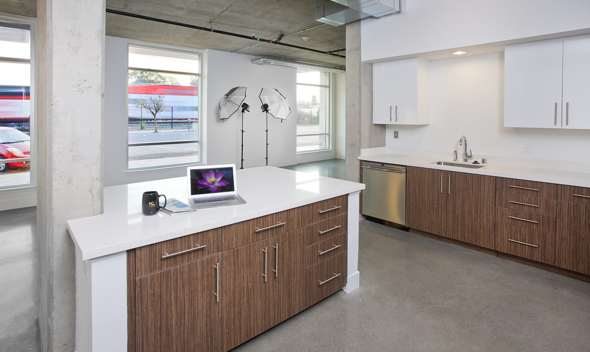 Kitchen of a loft unit with white and wood cabinetry, along with large windows viewing out to street.